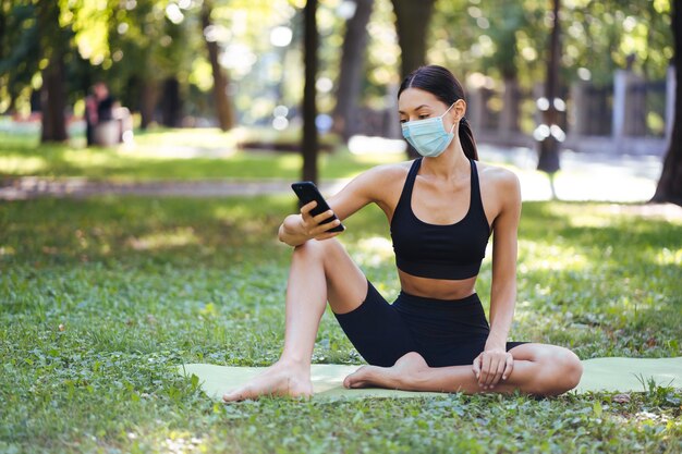 Fitness girl with a smartphone on nature background, enjoys sports training. Woman using cellphone outdoors.