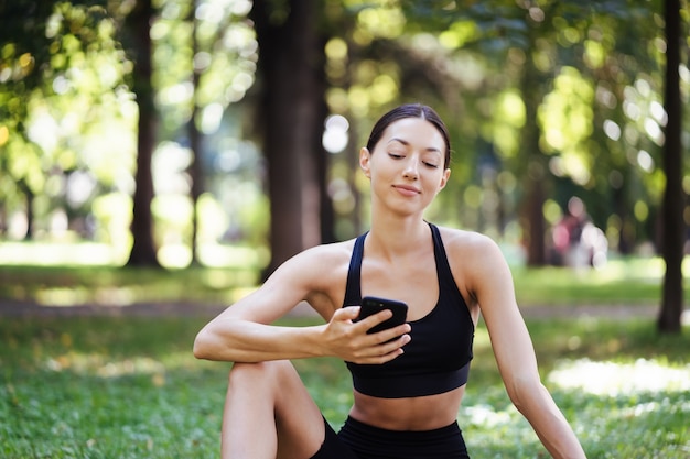 自然の背景にスマートフォンを持つフィットネスの女の子は、スポーツトレーニングを楽しんでいます。屋外で携帯電話を使用している女性。