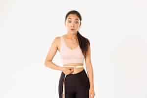 Free photo fitness, healthy lifestyle and wellbeing concept. surprised asian girl on diet, sportswoman wrap measuring tape around waist and look impressed as lose weight with workout.