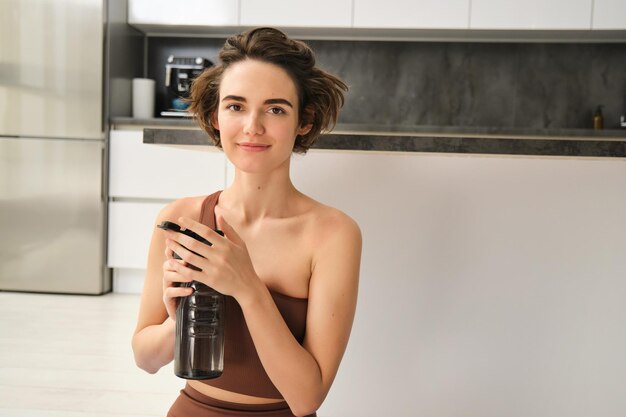 Fitness instructor smiling young woman workout at home sits on yoga mat and drinks water does exerci