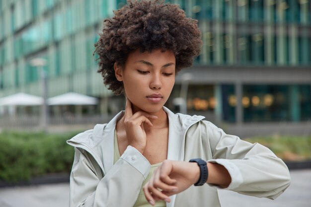 Fitness lady counts hearbeat checks pulse on neck concentrated at smartwatch monitors her health after workout dressed in sportswear poses outside against modern city building