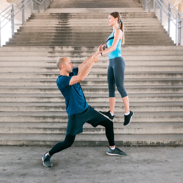 Fitness man lifting the young woman on his leg while practicing exercise near the staircase