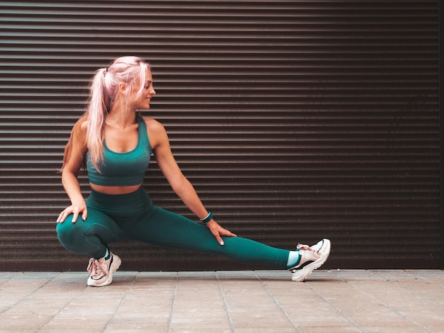 Fitness smiling woman in green sports clothing with pink hair Young beautiful model with perfect bodyFemale posing in the street near roller shutter wall Stretching out before training