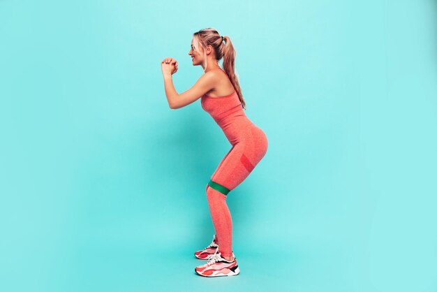 Fitness smiling woman in pink sports clothing Young beautiful model with perfect bodyFemale posing near blue wall in studioCheerful and happy Stretching out before training Doing squats