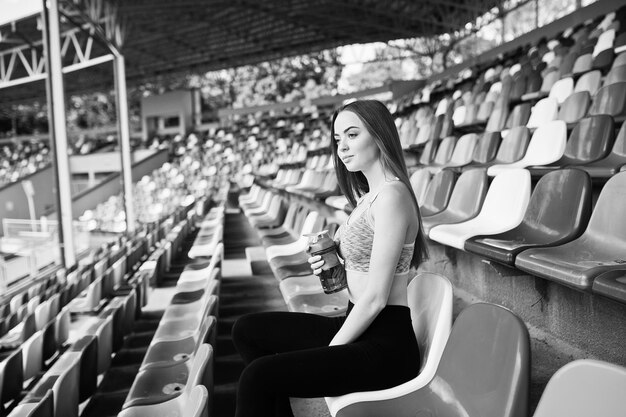 スタジアムの椅子に座っているスポーツウェアのフィットネススポーティな女の子屋外スポーツスポーツボトルのモックアップで幸せなセクシーな女性