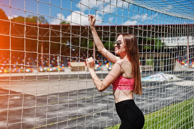 スタジアムのサッカーゲートでポーズをとったスポーツウェアとサングラスのフィットネススポーティな女の子屋外スポーツ幸せなセクシーな女性