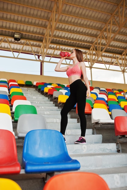 Foto gratuita fitness ragazza sportiva in abbigliamento sportivo e occhiali da sole poste allo stadio tribuna sport all'aria aperta donna sexy felice che beve acqua dalla bottiglia