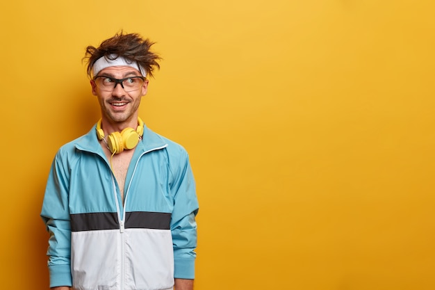 Fitness sporty guy in good mood, looks aside with motivation, enjoys workout and sport, listens music in headphones during training, dressed in active wear, copy space on yellow wall