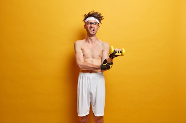 Fitness sporty guy raises heavy dumbbell, does exercise for arms, has much energy, feels pain, shows sport motivation, wears shorts and sport gloves. people, health, body care and home fitness concept