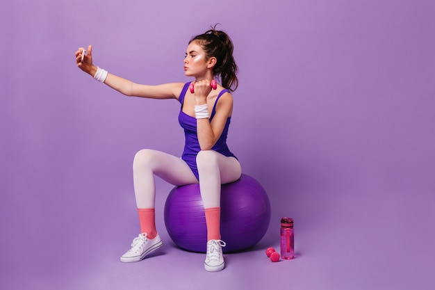 フィットネストレーナーの女性は、フィットボールに座って、ダンベルで運動し、紫色の壁で自分撮りをします