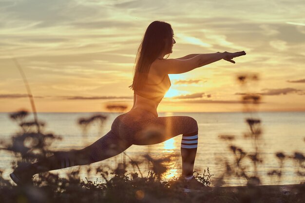 Fitness woman practicing stretching yoga exercises in nature against the background of the sea at sunset.