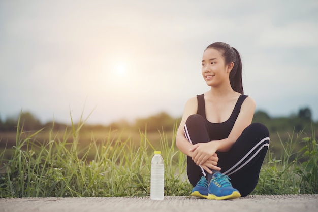  Fitness woman runner sit down relaxing with water bottle after training outside in park