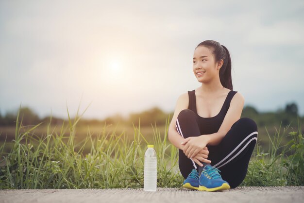  Fitness woman runner sit down relaxing with water bottle after training outside in park