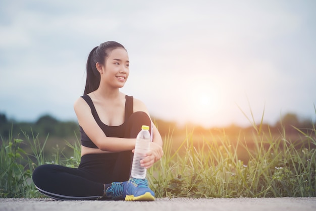  Fitness woman runner sit down relaxing with water bottle after training outside in park