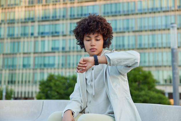 Fitness woman with curly hair reads message on smartwatch checks time listens music via wireless headphones dressed in sportsclothes poses against blurred building background has training outdoors