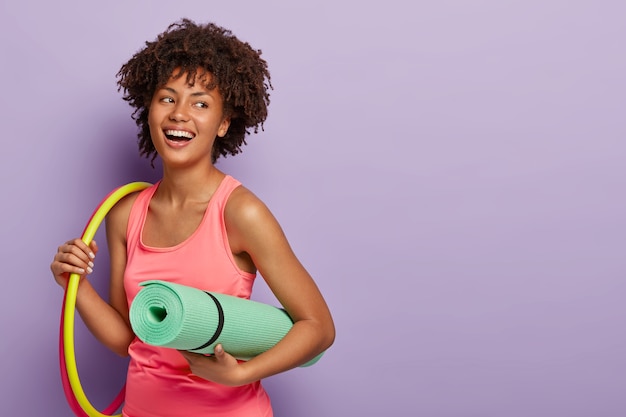 Fitness woman with dark skin, works with hula hoops, holds rolled karemat for training