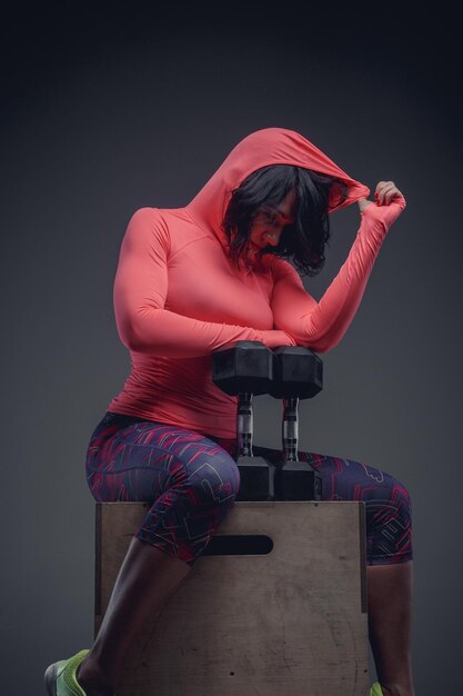 Fitness woman with short black hair in colorful sportswear sitting on wooden box with dumbell.