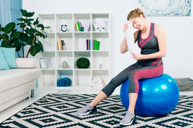 Fitness young woman sitting on blue pilates ball wiping sweat with towel