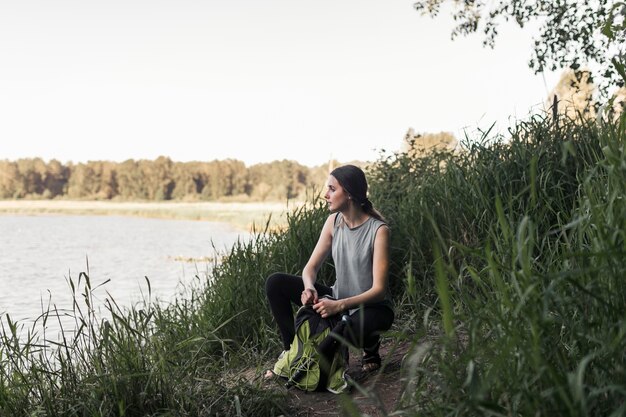 Фитнес молодая женщина с ее рюкзак, приседая возле озера