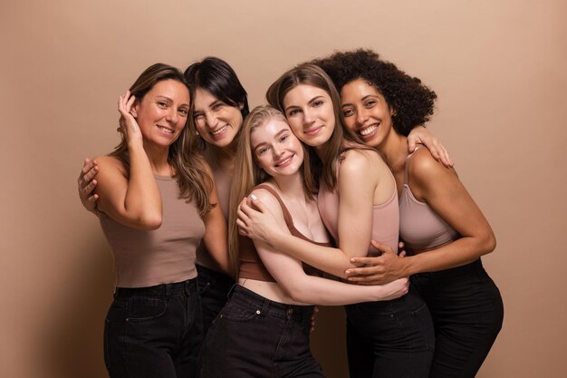 Five pretty african and caucasian women in brown tops black jeans look at camera on beige background