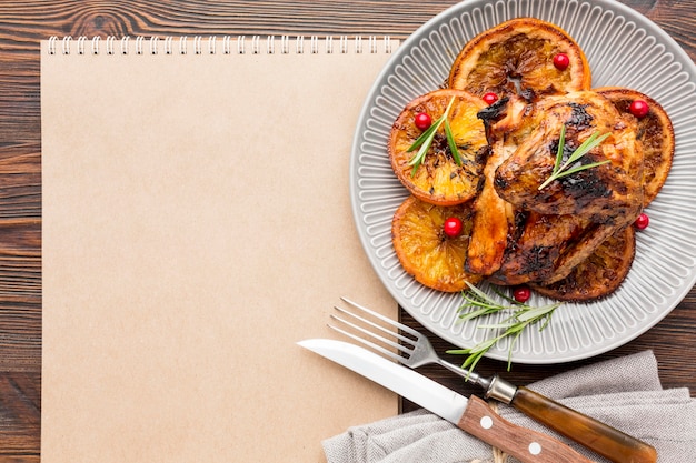 Flat lay baked chicken and orange slices on plate with cutlery and blank notepad