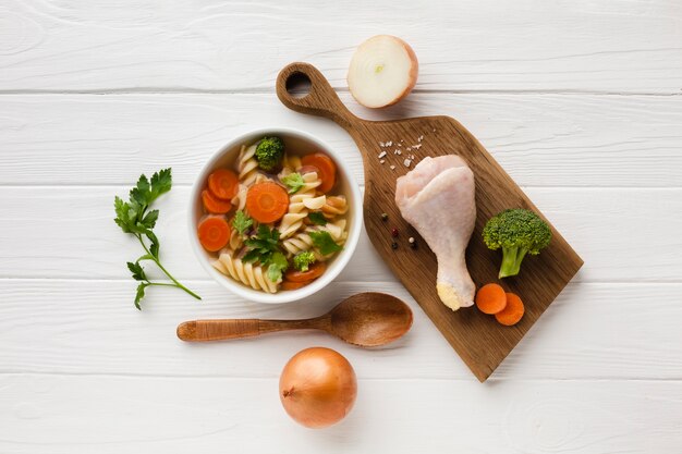 Flat lay broccoli carrots and fusilli in bowl with chicken drumstick on cutting board