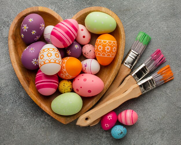 Flat lay of colorful easter eggs in heart shaped plate with paint brushes