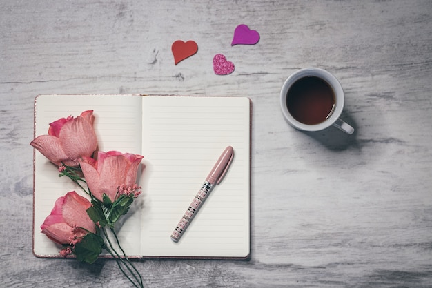 開いたノートブックの表面に一杯のコーヒー、柔らかいピンクの花、ペンを平らに置きます