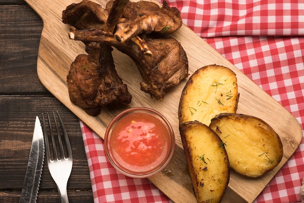 Flat lay lamb chops on wooden board with potatoes and sauce