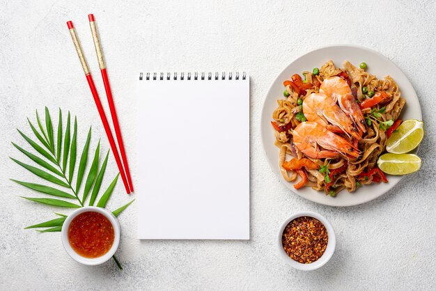 Flat lay noodles with vegetables and shrimp chopsticks and spices with blank notebook