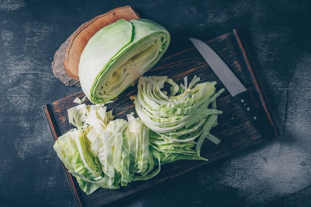 Flat lay sliced and chopped cabbage in cutting board and knife with wood stub on dark textured background. horizontal