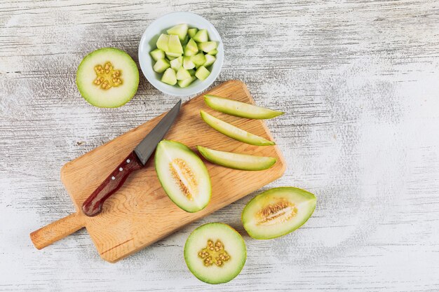 Flat lay sliced melon in wooden cutting board with melon in bowl and knife on white stone background. horizontal