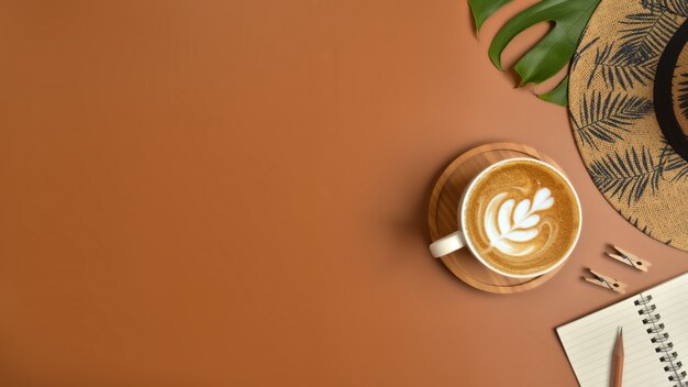 Flat lay, top view workspace with eye glasses, notebook, hat, pencil, green leaf, shoes and coffee cup on brown background.