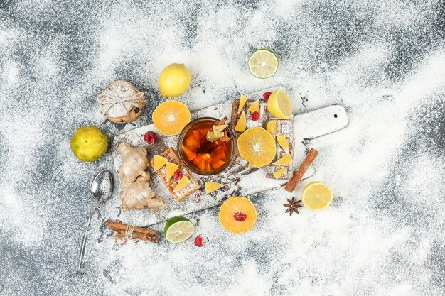 Flat lay waffles and rice wafers on white cutting board with herbal tea,citrus fruits,cinnamon and tea strainer on dark grey marble surface. horizontal