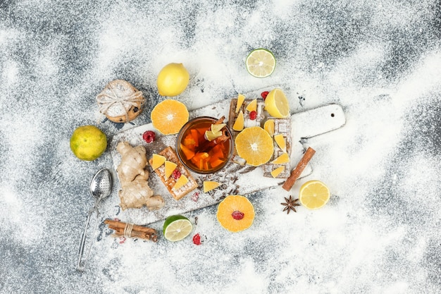 Flat lay waffles and rice wafers on white cutting board with herbal tea,citrus fruits,cinnamon and tea strainer on dark grey marble surface. horizontal