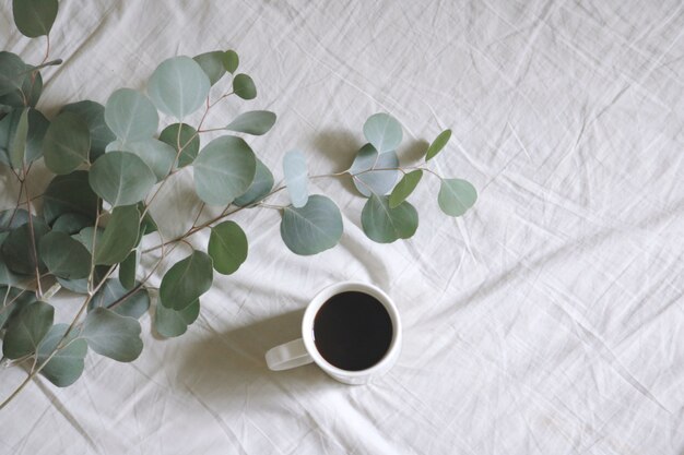 Плоская белая керамическая кружка с кофе рядом с серебряными листьями десен на белой простыне