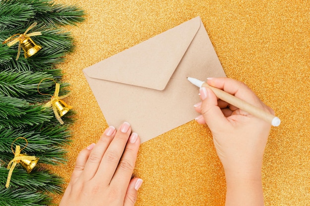 Flat lay on woman hands writing letter in santa claus craft envelope on golden shiny background with...