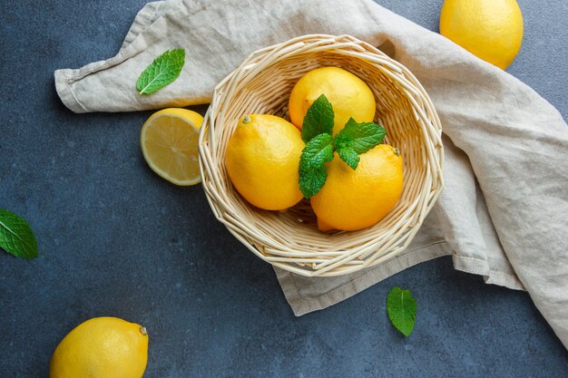 Flat lay yellow lemons and leaves in basket on white fabric cloth on dark surface. horizontal
