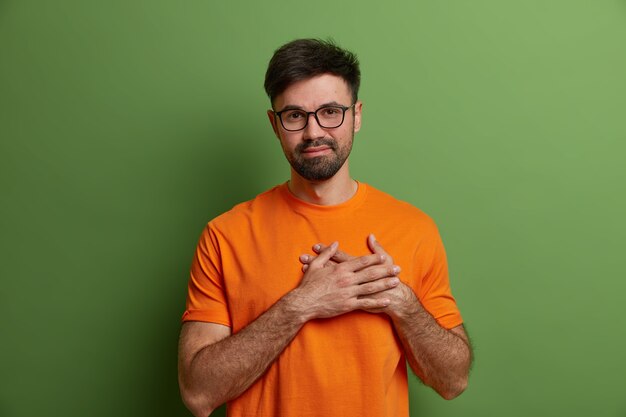 Flattered pleased unshaven guy presses hands to heart, expresses heartwarming feelings and gratitude appreciates praise wears transparent glasses and bright orange t shirt isolated on green wall