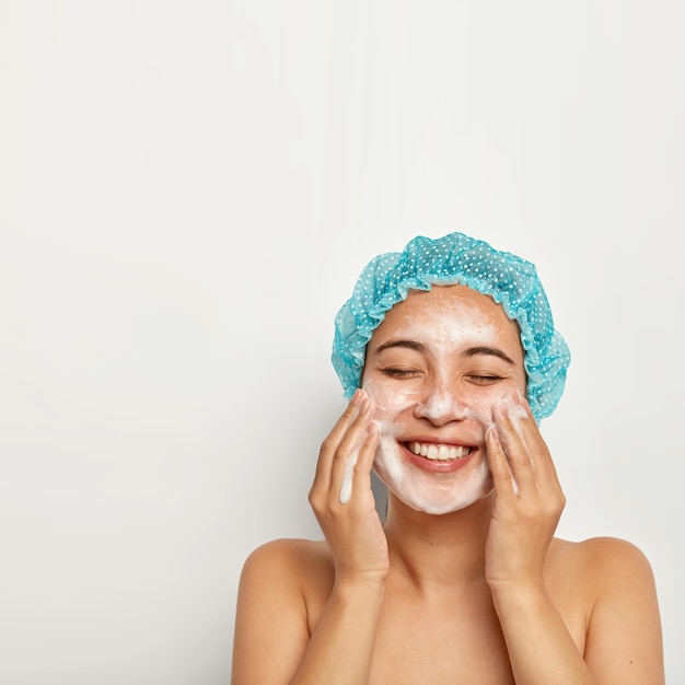 Flawless and purity of skin. Vertical image of pretty woman washes face, enjoys cold water, has foam on skin, smiles joyfully, keeps eyes closed, takes care of personal hygiene. Wellness concept