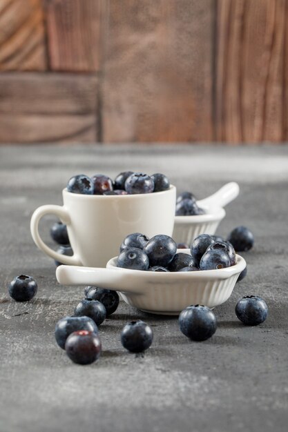 Fleshy blueberries in cup and ladles side view on grey surface