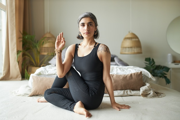 Flexible young advanced female yogi with premature gray hair sitting on floor in ardha matsyendrasana pose, doing seated spinal twist to improve digestion and relieve back pain