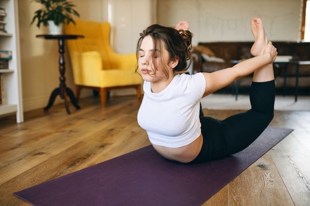 Flexible young female with curvy body exercising indoors, doing dhanurasana or bow pose on yoga mat to strenghten back, open chest and shoulders, improve digestion and stimulate reproductive organs