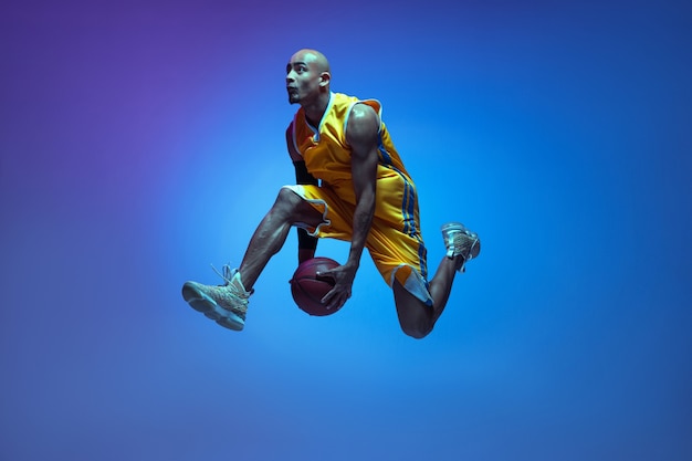 Volo. bello giocatore di basket maschio afroamericano in movimento e azione alla luce al neon sulla parete blu.