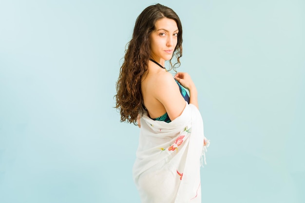 Free photo flirting with you. charming caucasian woman wrapped in a beach sarong while looking at the camera with a flirty look against a light background