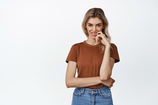 Flirty blond girl looking at amera with smiling cunning face expression has an idea standing against white background