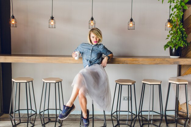 青い目とコーヒーショップでコーヒーを飲みながら椅子に座っている明るいピンクの唇を持つ軽薄なブロンドの女の子。