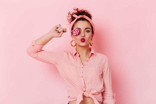 Кокетливая темноволосая девушка в розовой повязке на голову и массивных серьгах прикрывает глаз конфетой.