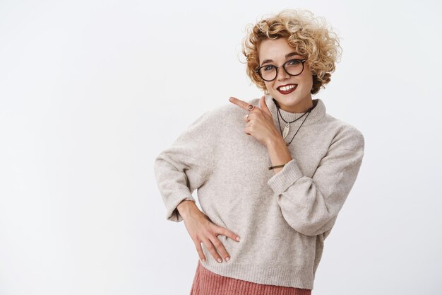 Flirty and energized excited cute girlfriend with blond short haircut and glasses in sweater pointing left and smiling broadly, looking happy and friendly.