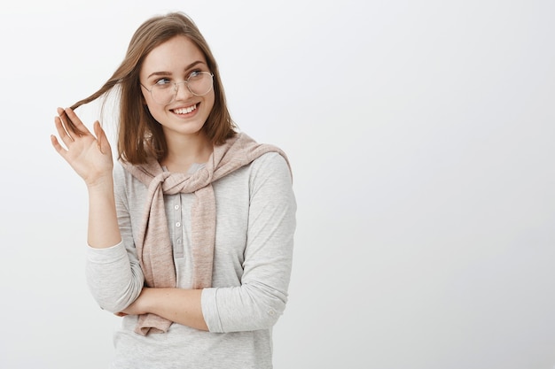 Flirty feminine charming european female in glasses and sweatshirt tied over neck playing with strand of hair smiling and glancing right with admiration and desire posing over white wall